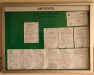 Ein Bulletinbrett mit grünem Hintergrund, beschriftet mit 'AMTSTAFEL'. Es sind mehrere weiße Blätter mit gedrucktem Text befestigt, in einem Rastermuster angeordnet. Die Blätter scheinen Jobangebote oder Ankündigungen zu enthalten.