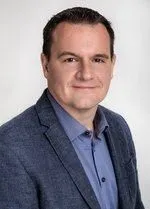 Ein Mann lächelt und posiert für ein Porträt. Er trägt ein blaues Hemd und einen blauen Blazer. Er hat kurze braune Haare und einen hellen Teint. Der Hintergrund ist weiß.