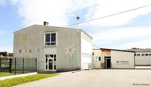 Ein Kindergartengebäude mit einem grünen Zaun, einer weißen Tür und einer Blumenwand. Es ist in Weiß und Grau gestrichen, mit einem Schornstein auf dem Dach.