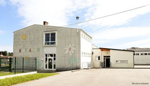 Ein Kindergartengebäude mit einem grünen Zaun, einer weißen Tür und einer Blumenwand. Es ist in Weiß und Grau gestrichen, mit einem Schornstein auf dem Dach.