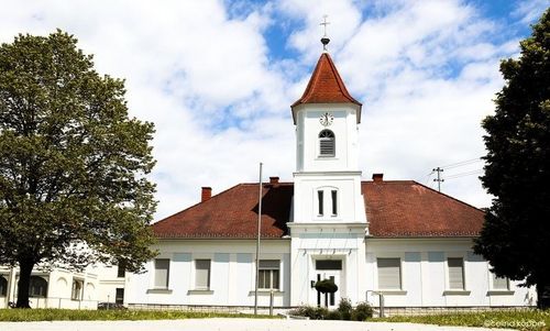 Ein weißes Gebäude mit rotem Dach und einem Turm mit Uhr steht in einem Rasenbereich mit Bäumen und Stromleitungen.