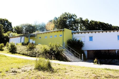 Ein modernes Gebäude mit gelber Fassade befindet sich auf einem Hügel. Es verfügt über mehrere Fenster und eine Metalltreppe, die zum Eingang führt. Das Gebäude ist von üppigem Grün und Bäumen umgeben.