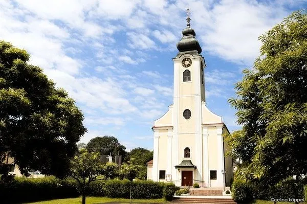 Eine weiße Kirche mit einem Turm und einer Uhr. Sie ist von Bäumen und Büschen umgeben.