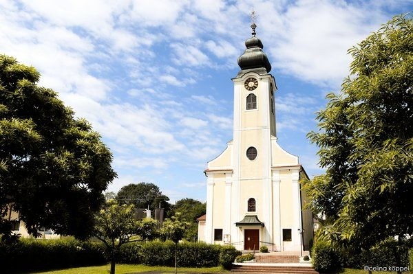 Eine weiße Kirche mit einem Turm und einer Uhr. Sie ist von Bäumen und Büschen umgeben.