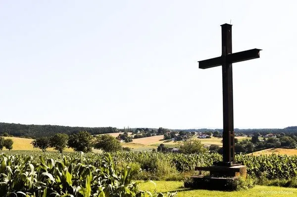 Ein großes Metallkreuz steht inmitten eines Maisfeldes mit einem Dorf im Hintergrund.