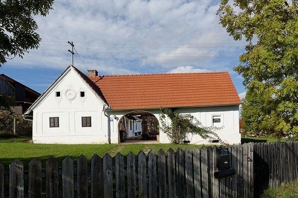 Ein weißes Haus mit einem roten Dach steht auf einem Rasen und ist von einem Holzzaun umgeben. Bäume und Grünpflanzen sind im Hintergrund zu sehen.