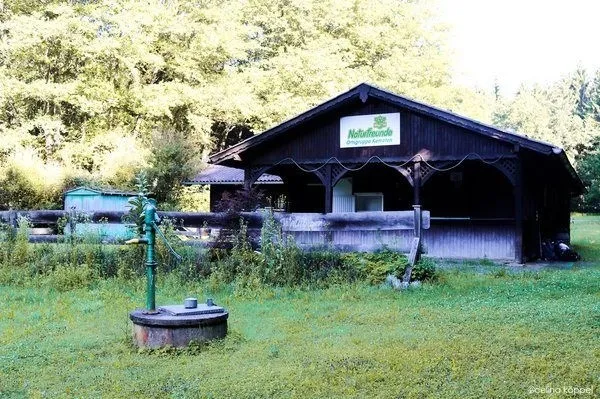 Ein altes Holzgebäude mit einem Schild, Naturfreunde, steht auf einem Feld. Es hat ein geneigtes Dach und Holzzäune. Im Vordergrund befindet sich eine grüne Pumpe.