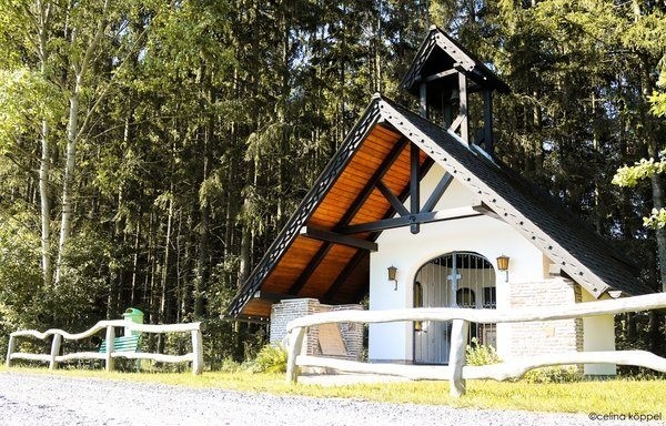 Eine kleine Kapelle mit einem Holzzaun, einer grünen Bank und einem Mülleimer im Wald.
