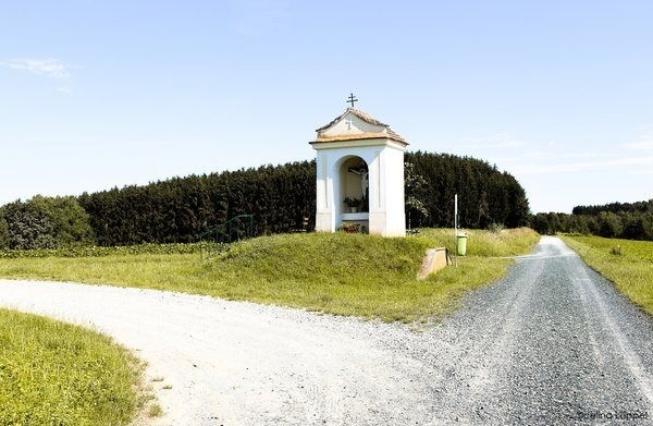 Eine kleine Kapelle mit einem Kreuz auf der Spitze steht auf einem grasbewachsenen Hügel mit Kieswegen auf beiden Seiten. Im Hintergrund sind Bäume und ein klarer blauer Himmel zu sehen.