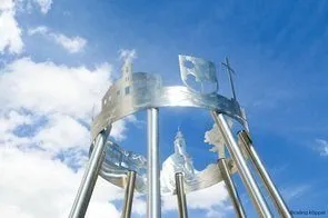 Ein Nahaufnahme einer Windglocke aus Edelstahl, die verschiedene architektonische und religiöse Symbole zeigt. Die Windglocke ist vor einem klaren blauen Himmel mit verstreuten Wolken zu sehen.