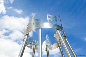 Ein Nahaufnahme einer Windglocke aus Edelstahl, die verschiedene architektonische und religiöse Symbole zeigt. Die Windglocke ist vor einem klaren blauen Himmel mit verstreuten Wolken zu sehen.