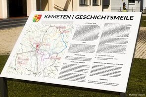 Ein Schild mit Karte und Text steht auf Gras und zeigt 'Kemeten | Geschichtsmeile'. Die Karte markiert Orte mit roten und blauen Punkten. Der Text handelt von historischen Stätten und ist vor weißem Hintergrund.