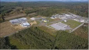 Luftaufnahme eines Industriegebiets, umgeben von dichtem Baumbestand und Vegetation. Gebäude und Strukturen sind sichtbar, zusammen mit einer Straße, die zum Gebiet führt.