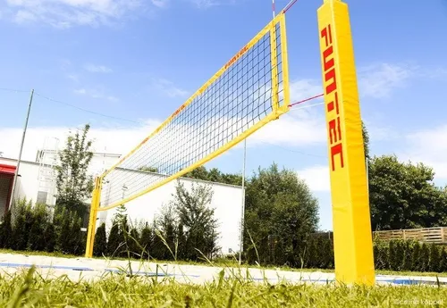 Ein gelbes FUNTEC Volleyballnetz steht auf einem Rasenfeld mit einem weißen Gebäude im Hintergrund. Das Netz ist an gelben Stangen befestigt und steht in der Mitte eines sonnigen Tages.