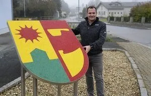 Ein Mann steht neben einem farbenfrohen Schild auf einem Kiesweg, in der Nähe einer Straße mit Häusern und Bäumen.