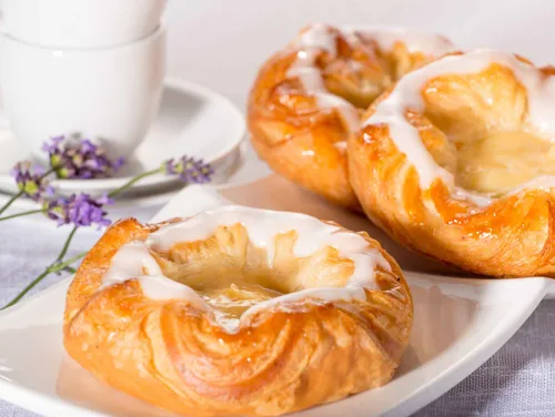 Drei Gebäckstücke mit weißer Glasur auf einem weißen Teller, mit einer weißen Tasse und Untertasse, und Lavendelblüten im Hintergrund.