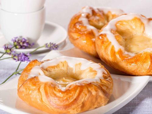 Drei Gebäckstücke mit weißer Glasur auf einem weißen Teller, mit einer weißen Tasse und Untertasse, und Lavendelblüten im Hintergrund.
