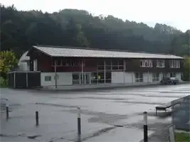 Ein großes Gebäude mit einem Metalldach und vielen Fenstern befindet sich in einem ländlichen Gebiet mit Bergen im Hintergrund. Der vordere Parkplatz ist nass und leer.