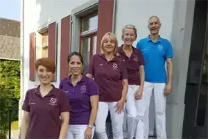 Fünf Personen stehen vor einem Gebäude mit roten Fensterläden. Sie tragen alle lila Hemden und weiße Hosen, außer der Mann rechts, der ein blaues Hemd trägt.