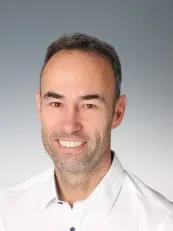 Ein Mann mit kurzem Bart, dunklem Haar und weißem Hemdkragen lächelt in die Kamera. Er steht vor grauem Hintergrund.