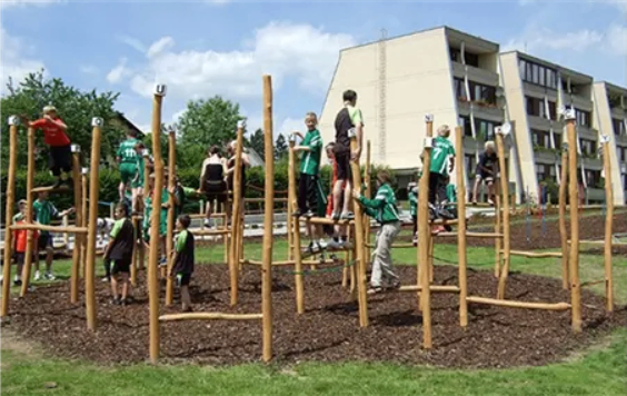 Eine Gruppe von Kindern klettert auf einem hölzernen Hindernisparcours in einem Park mit Gebäuden im Hintergrund.