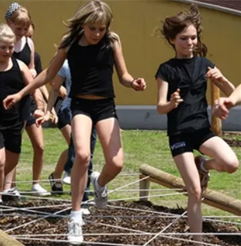 Eine Gruppe von Kindern in schwarzen T-Shirts und Shorts nimmt an einem Hindernisparcours teil, wobei ein Mädchen über einen Baumstamm springt.
