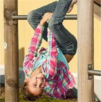 Ein junges Mädchen hängt kopfüber an einer Spielplatzstange, lächelt und trägt ein rosa-weißes kariertes Hemd und Jeans.