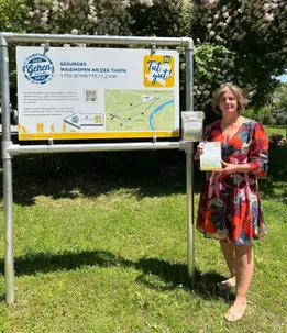 Eine Frau steht vor einem Wegweiser mit Karte und hält ein Blatt Papier in der Hand. Der Schild sagt 'Gehen' und 'Gesundes Waidhofen an der Thaya'. Im Hintergrund sind Bäume und ein Weg zu sehen.