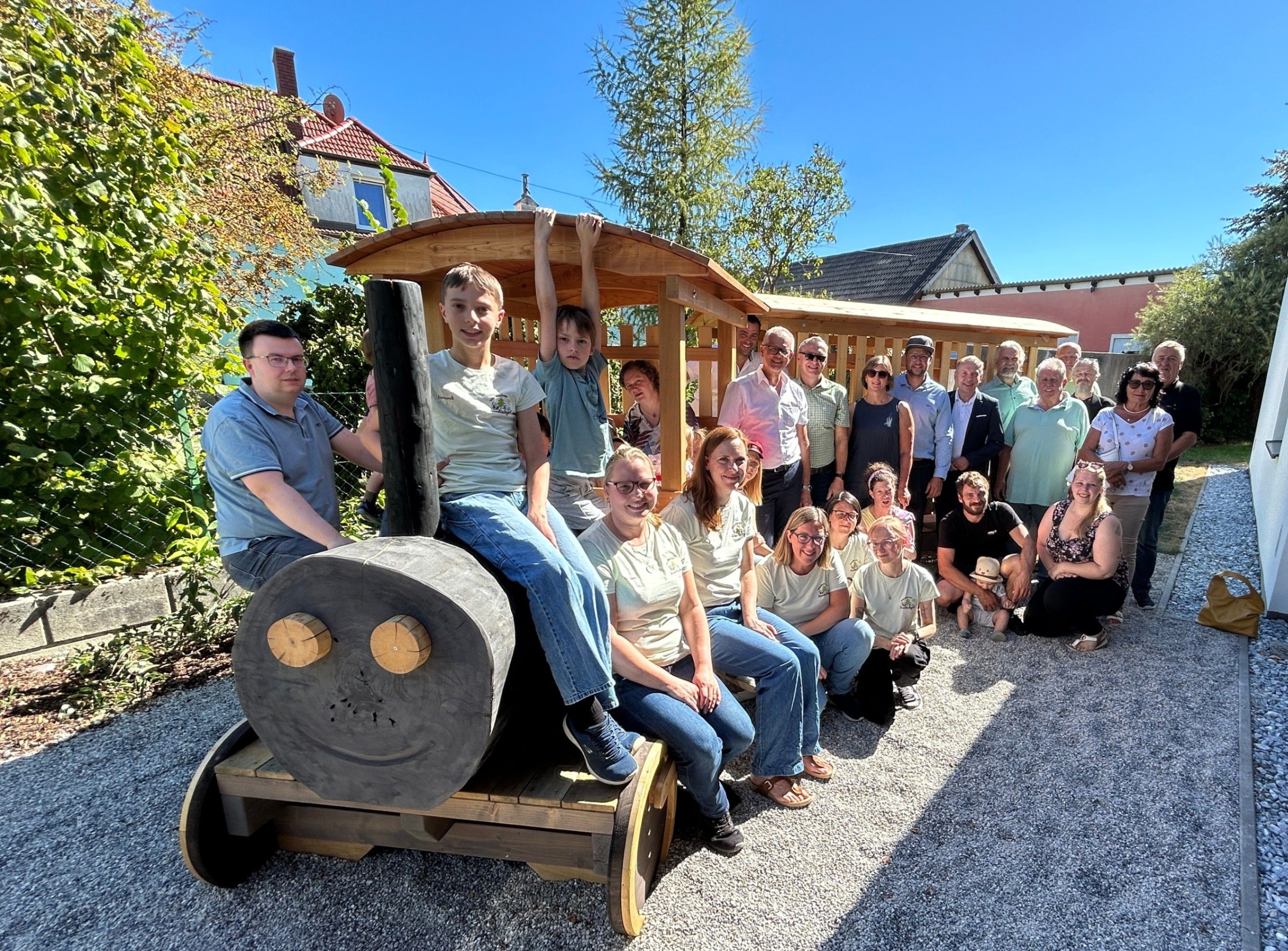 Eine Gruppe von Menschen, einschließlich Kindern, posiert für ein Foto neben einer Holzbahn in einer Gartenumgebung. Die Erwachsenen tragen Freizeitkleidung, und die Kinder sitzen auf der Bahn.