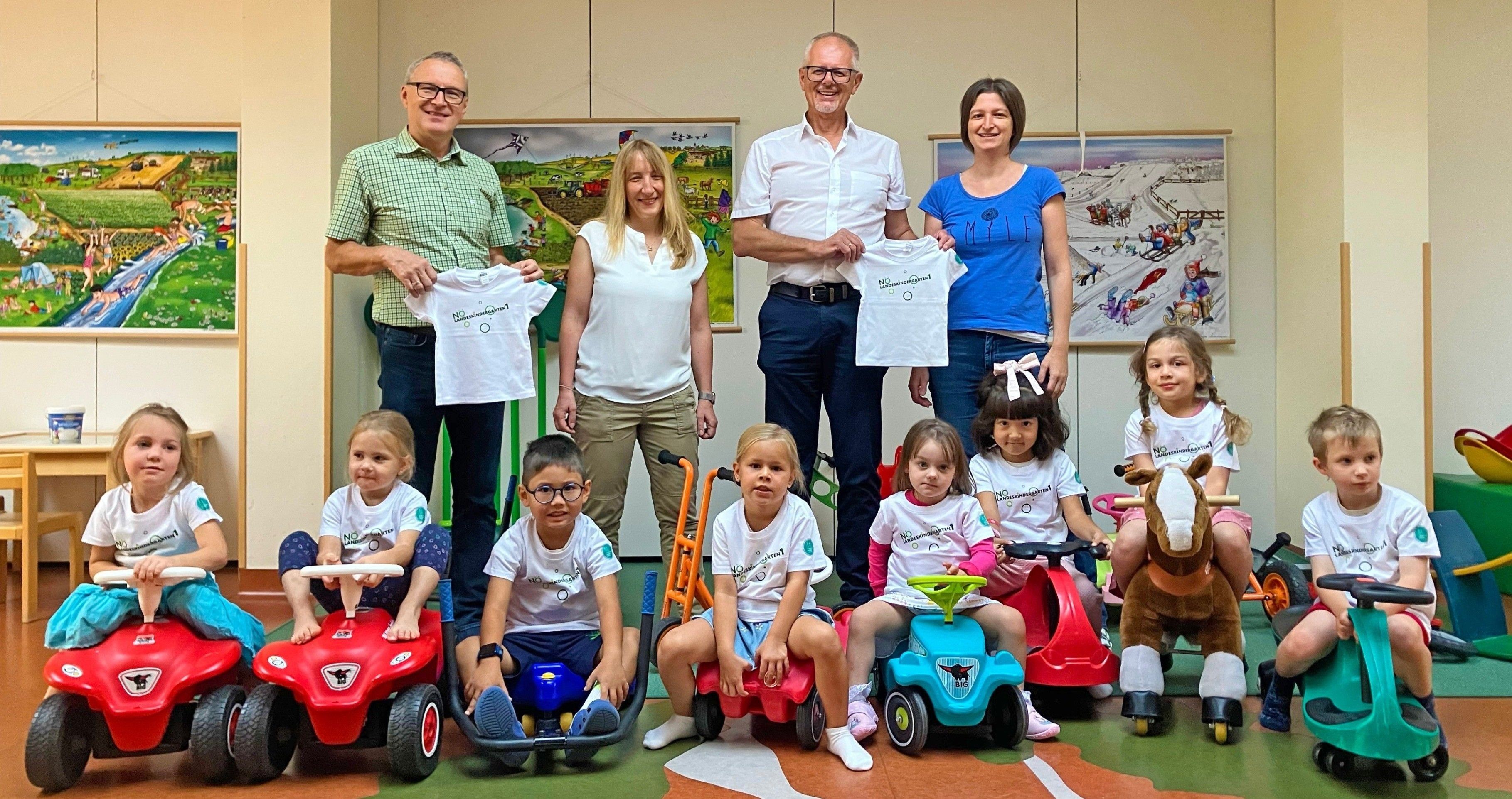 Mehrere Erwachsene und Kinder posieren für ein Bild mit weißen T-Shirts und Spielzeug, zwei Erwachsene halten weiße T-Shirts mit Designs, ein Kind sitzt auf einem roten Spielzeugauto und zwei Mädchen sitzen auf blauen Spielzeugautos.