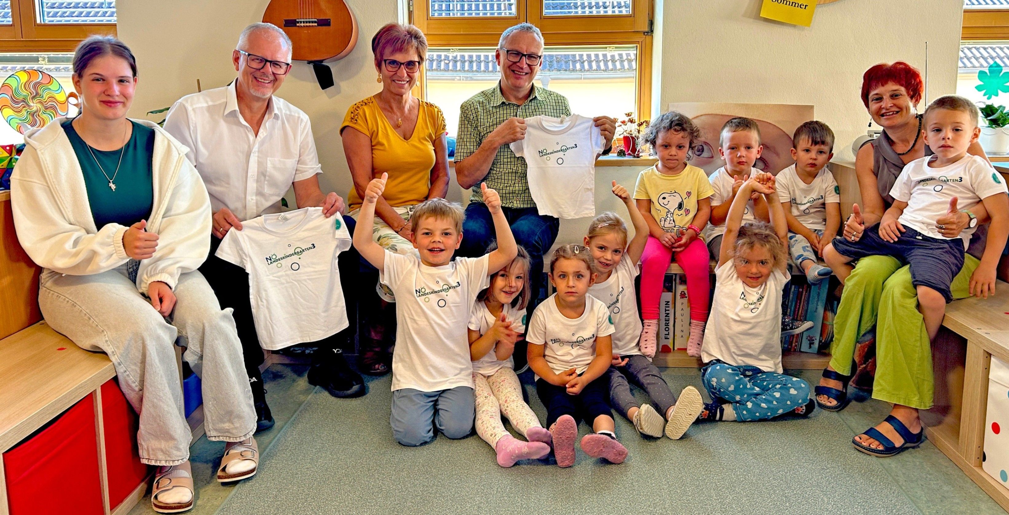 Eine Gruppe von Erwachsenen und Kindern sitzt und hockt auf dem Boden und lächelt. Zwei Erwachsene halten weiße T-Shirts mit Logos hoch, während andere ihre Shirts hochhalten. Eine Gitarre und Bücher sind im Hintergrund.