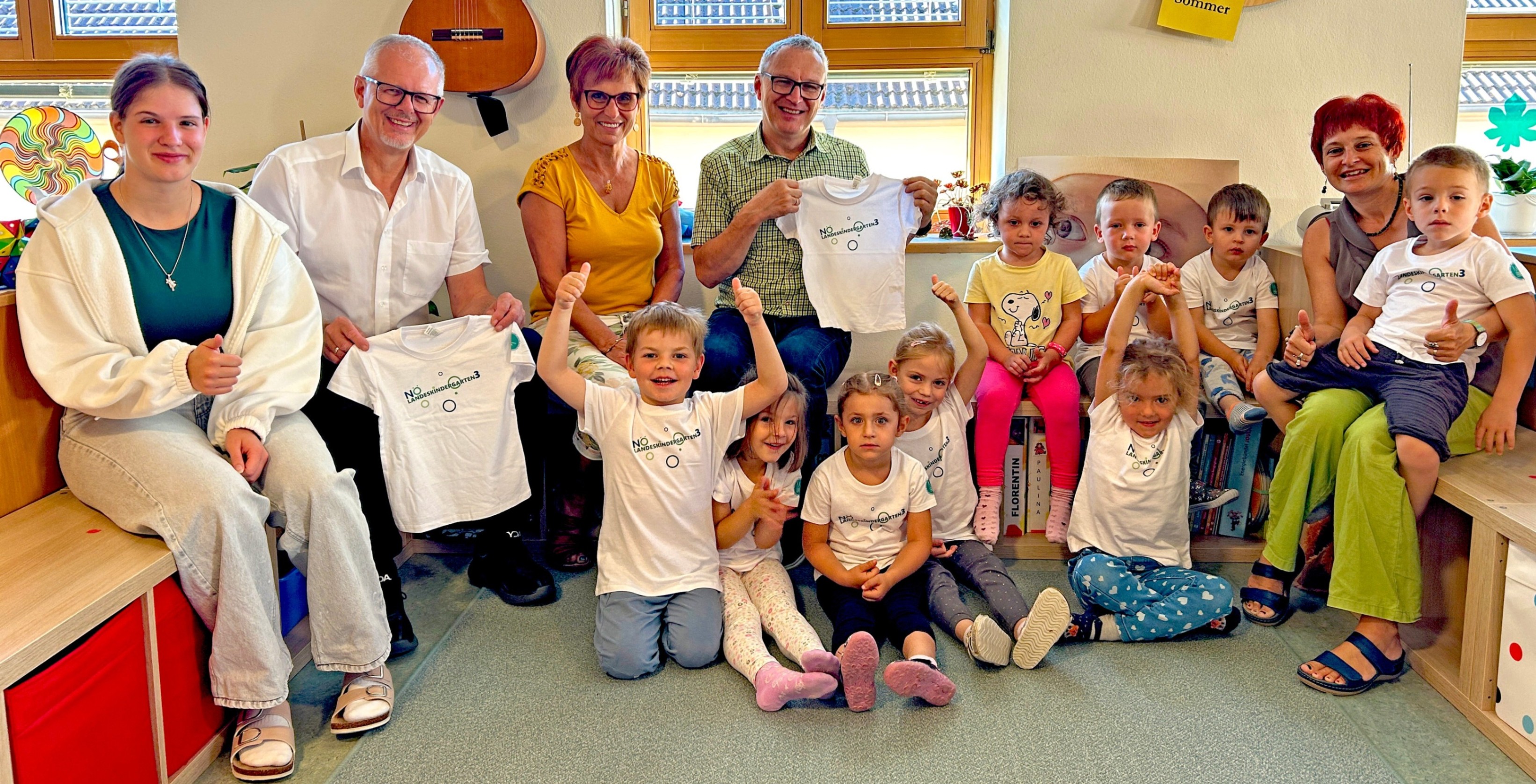 Eine Gruppe von Erwachsenen und Kindern sitzt und hockt auf dem Boden und lächelt. Zwei Erwachsene halten weiße T-Shirts mit Logos hoch, während andere ihre Shirts hochhalten. Eine Gitarre und Bücher sind im Hintergrund.