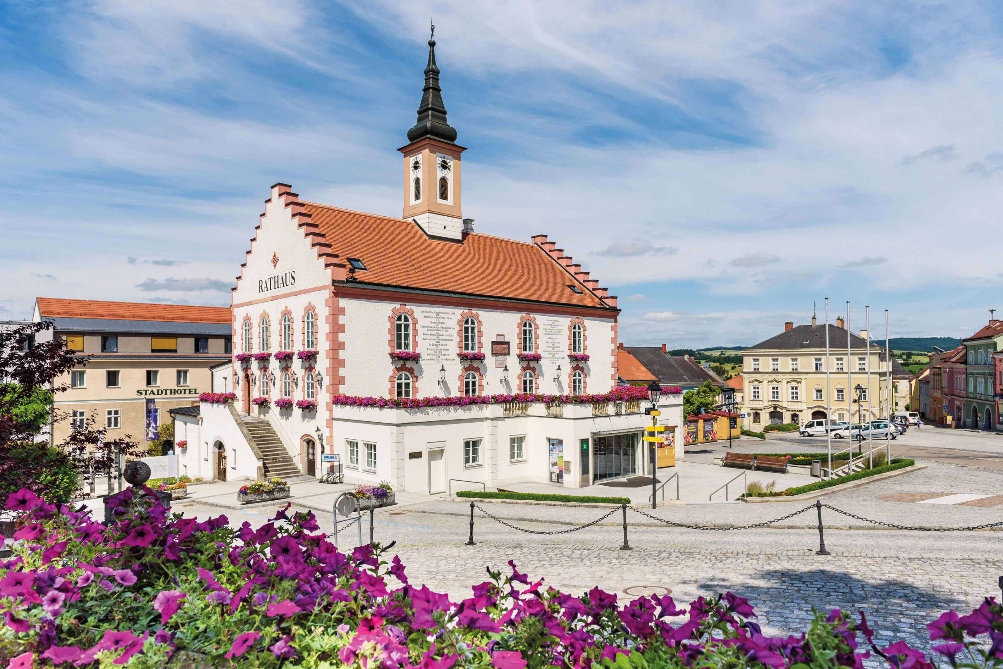 Das Rathaus, ein weißer und roter Bau mit einem Turm, ist mit Blumen geschmückt. Es befindet sich auf einem Stadtplatz mit Kopfsteinpflaster und ist von grünen Pflanzen umgeben.