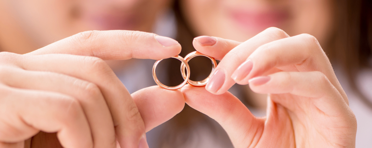 Zwei goldene Eheringe werden in den Fingern einer Frau gehalten. Der Fokus liegt auf den Ringen, wobei das Gesicht der Frau im Hintergrund verschwommen ist.