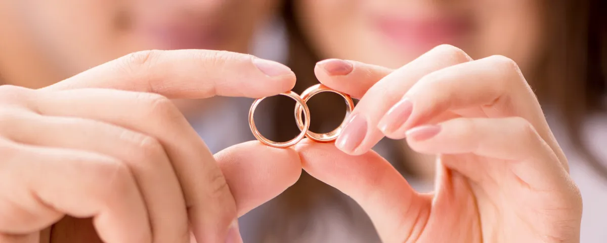 Zwei goldene Eheringe werden in den Fingern einer Frau gehalten. Der Fokus liegt auf den Ringen, wobei das Gesicht der Frau im Hintergrund verschwommen ist.