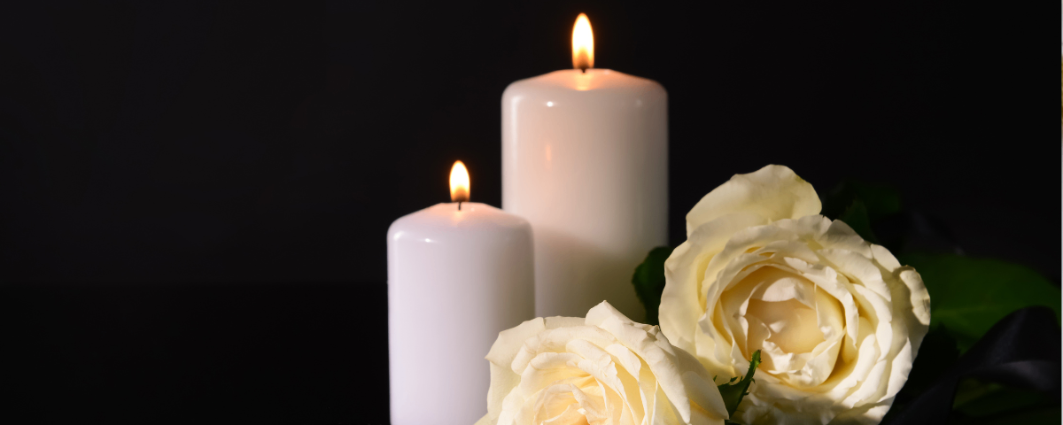 Zwei weiße Kerzen sind vor einem dunklen Hintergrund angezündet, mit weißen Rosen im Vordergrund.