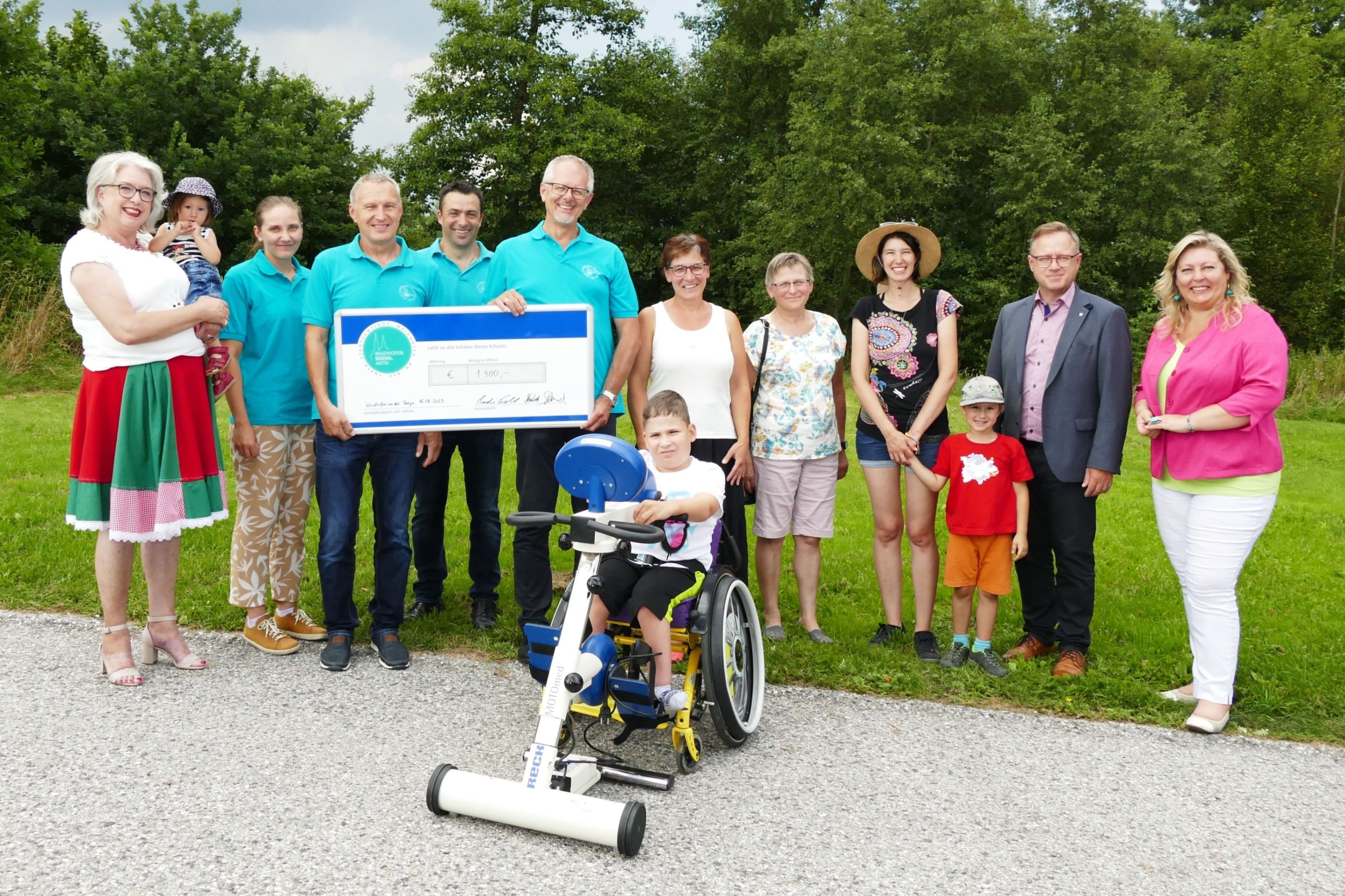 Eine Gruppe von Menschen, darunter Erwachsene und Kinder, steht um ein Kind im Rollstuhl herum und hält einen großen Scheck. Sie befinden sich in einem Grasbereich mit Bäumen im Hintergrund.