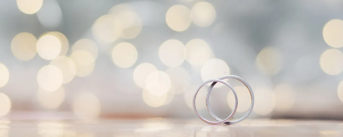 Zwei silberne Eheringe sind vor einem unscharfen Hintergrund mit warmen Lichtern dargestellt. Die Ringe liegen eng beieinander und symbolisieren Einheit und Zusammengehörigkeit.