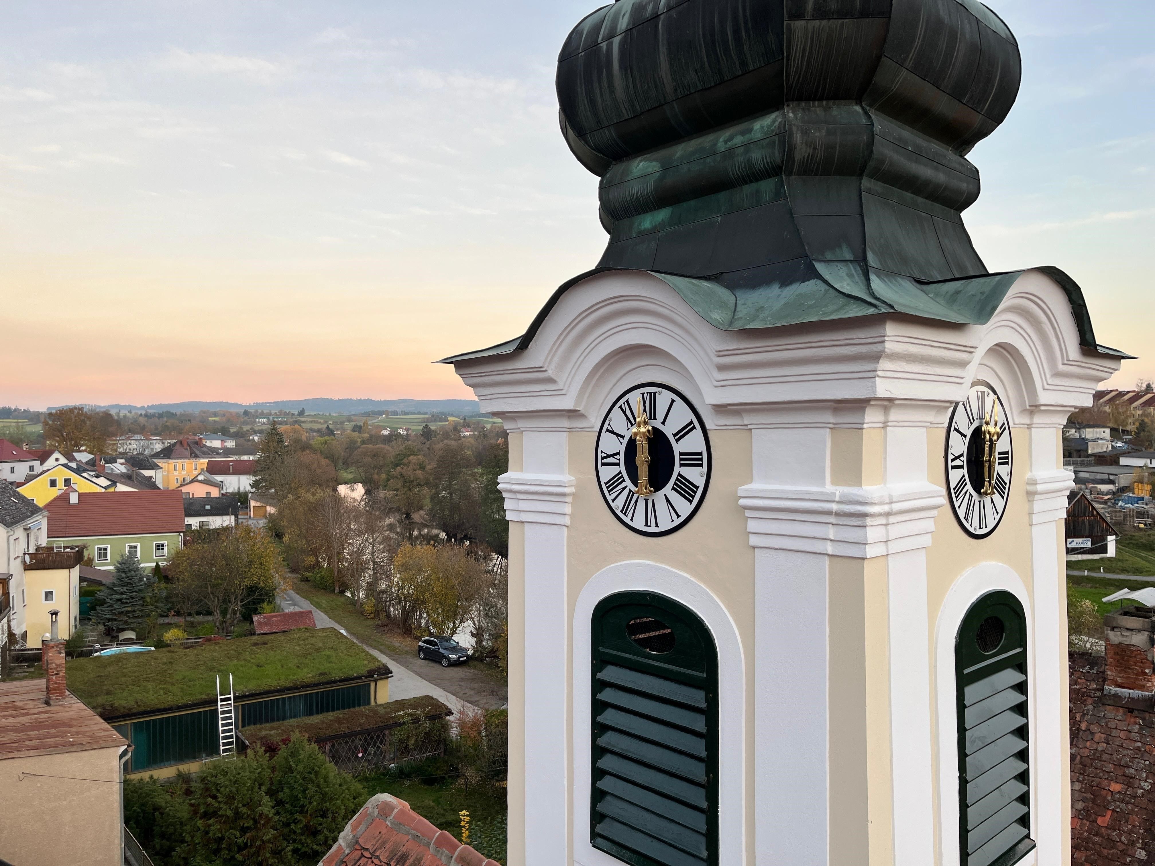 Ein Turm mit zwei Uhren, eine zeigt 10:35, steht auf einem Dach mit einer grünen Kuppel. Dahinter ist eine Stadtlandschaft unter einem teilweise bewölkten Himmel zu sehen.