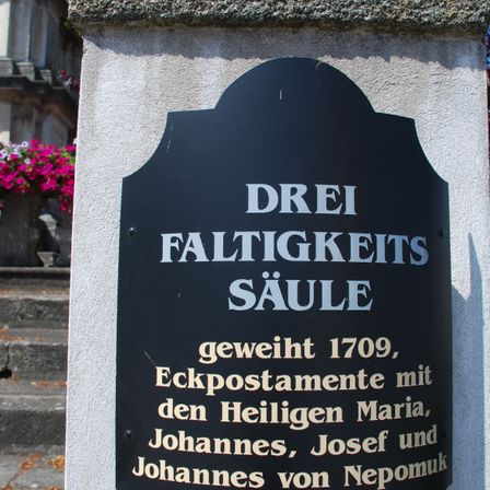 Eine schwarze Plakette an einer Wand trägt den weißen Schriftzug 'Drei Faltigkeits Saule', was 'Dreieinigkeitssäule' bedeutet. Sie ist den Heiligen Maria, Johannes, Josef und Johannes von Nepomuk gewidmet. Sie wurde 1709 geweiht.