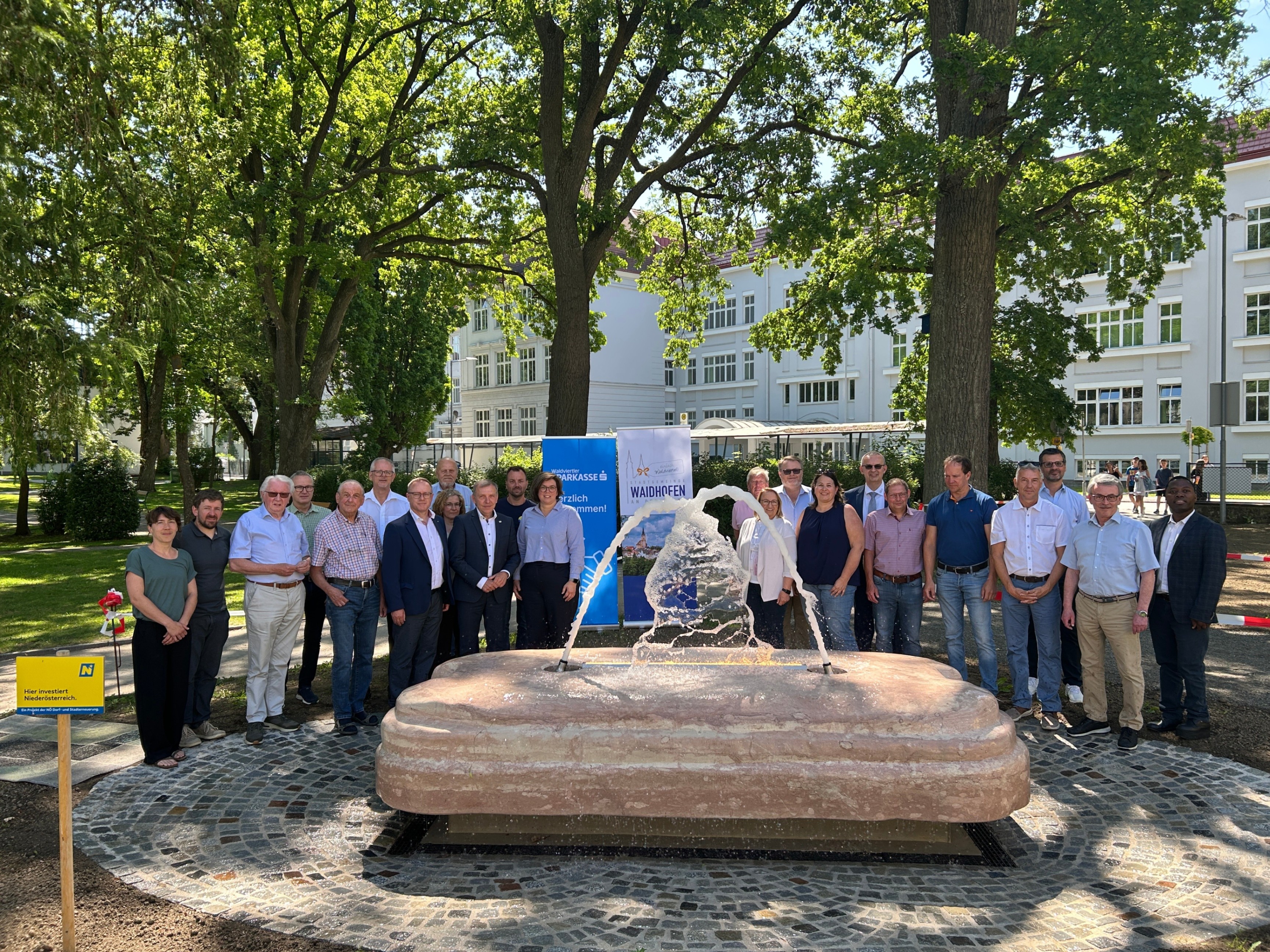 Eine Gruppe von Menschen in Geschäftskleidung versammelt sich um einen Brunnen, mit einem großen Gebäude und Bäumen im Hintergrund.