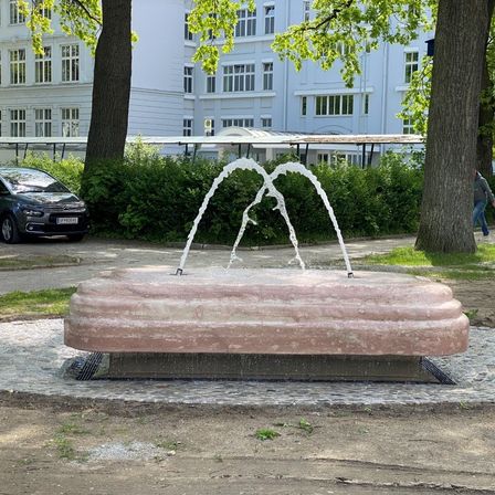 Ein Springbrunnen sprüht Wasser in die Luft, mit einer rosa Steinbasis, umgeben von Bäumen und Büschen. Ein Auto ist in der Nähe geparkt. Eine Person geht auf dem Bürgersteig im Hintergrund.