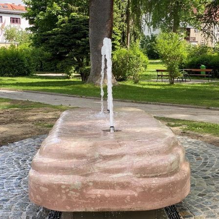 Ein Brunnen mit Wasserstrahl in der Mitte eines Parks. Bäume und eine Bank im Hintergrund.