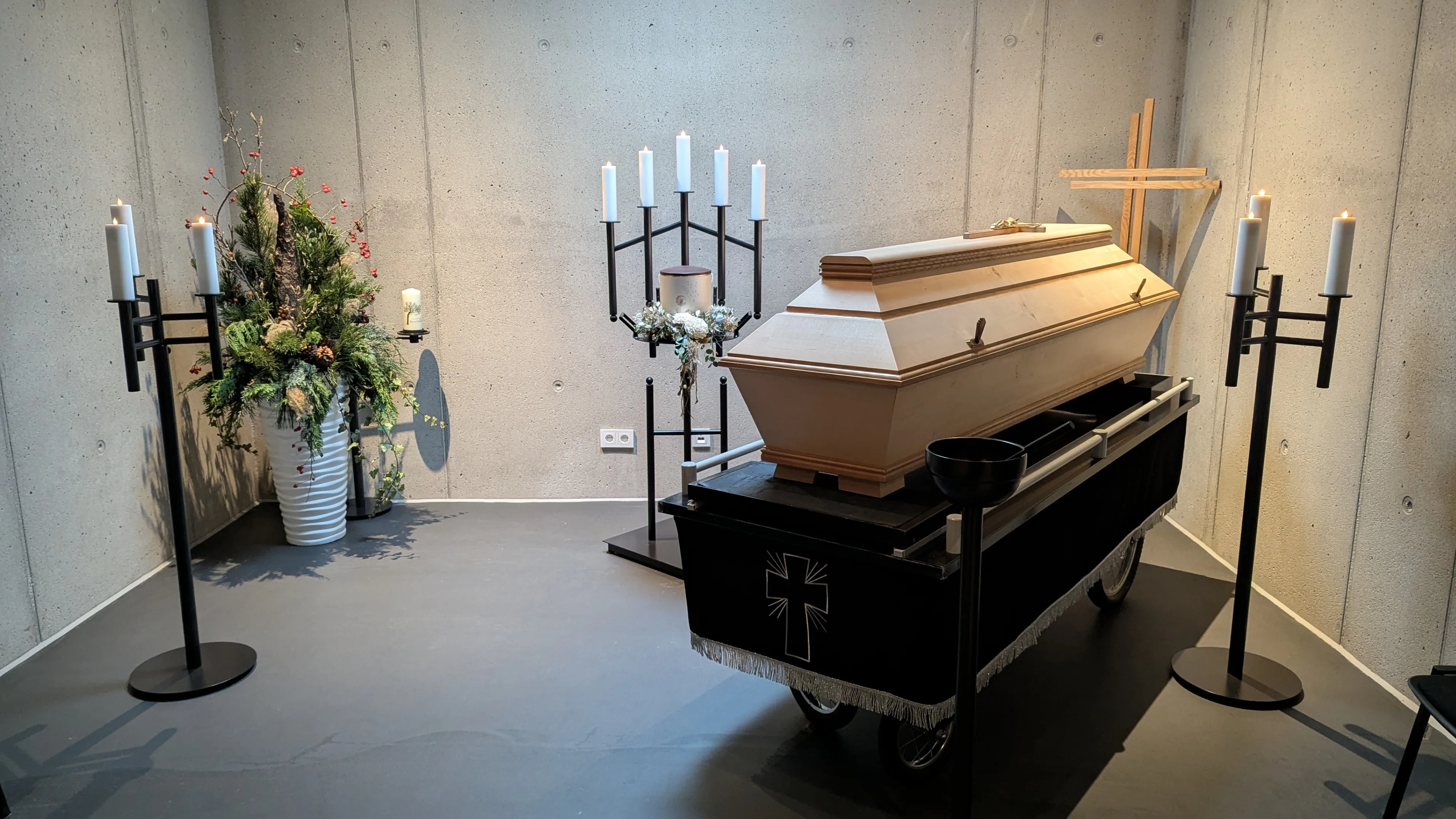 Ein Sargwagen mit einem Sarg steht in einem Raum, begleitet von einem Kerzenständer und einem Kreuz.