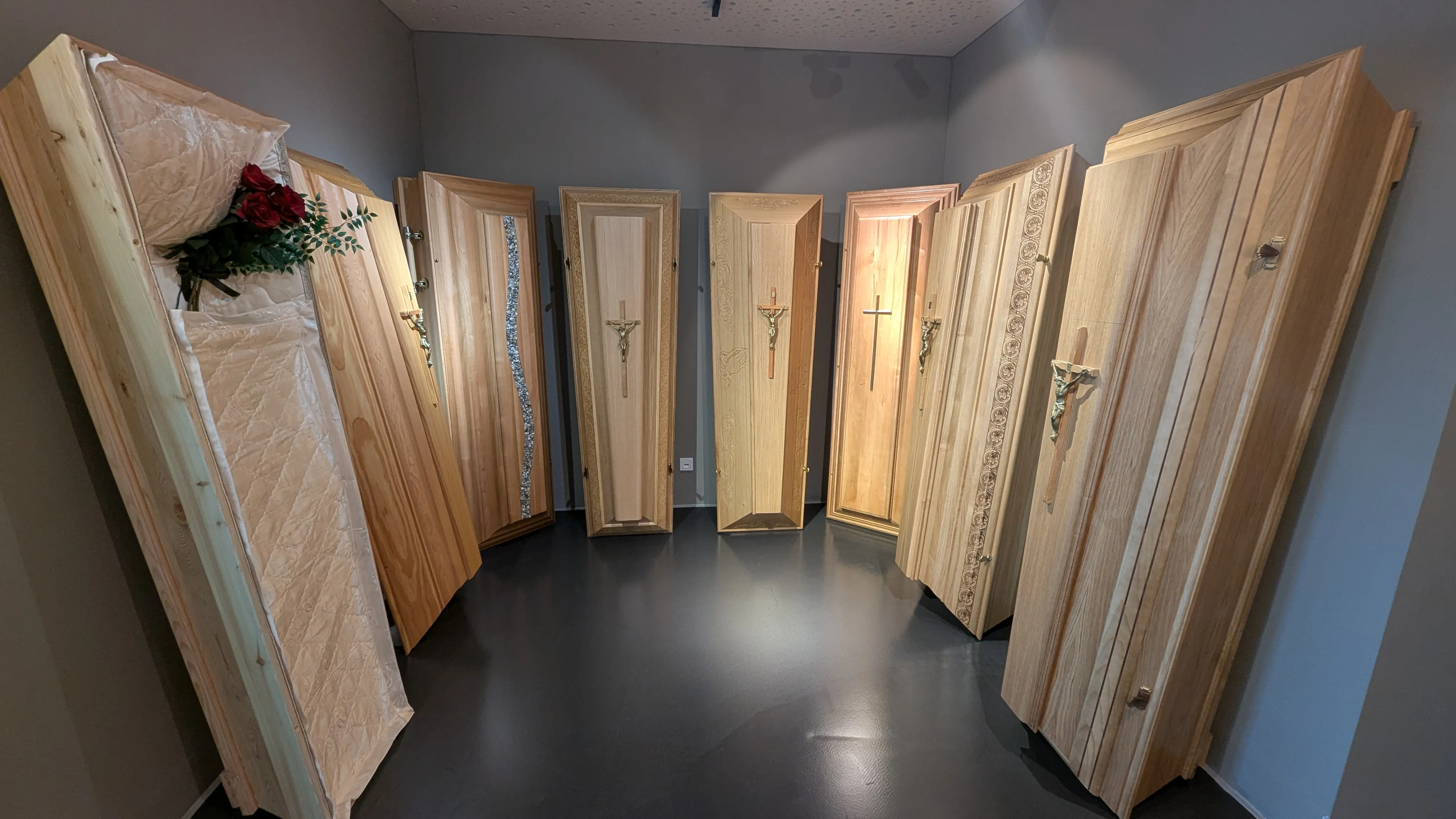 Ein Raum zeigt mehrere Holzsärge mit kunstvollen Designs, einschließlich Kreuzen, die an einer grauen Wand angeordnet sind.