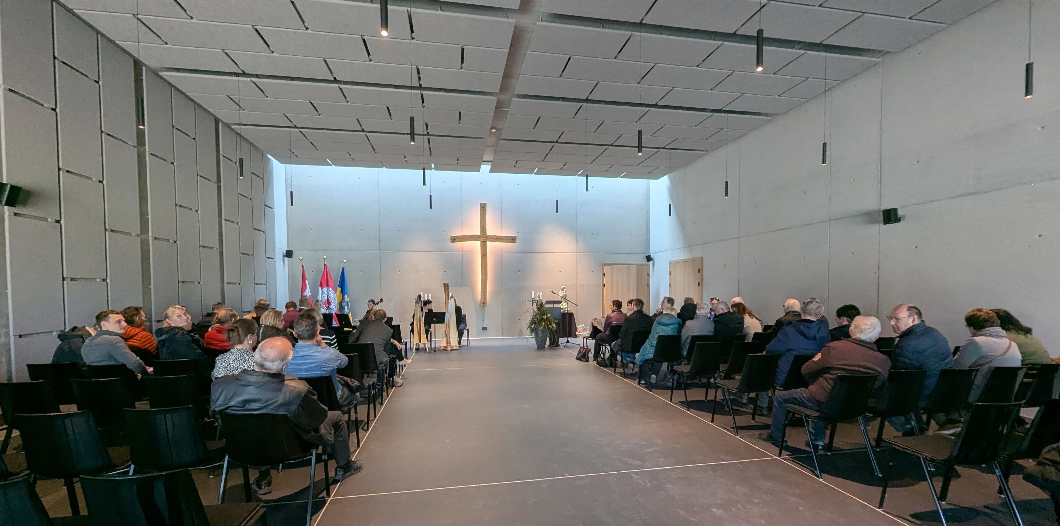 Ein Raum, der einer Kapelle ähnelt, mit einem hölzernen Kreuz an der Wand, einer Gruppe sitzender Personen, einer Person, die Harfe spielt, und Flaggen auf der linken Seite.
