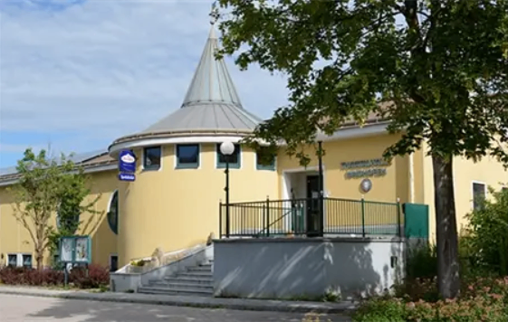 Ein modernes gelbes Gebäude mit einem kegelförmigen Dach und großen Fenstern ist an einem sonnigen Tag zu sehen.