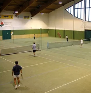 Ein Hallentennisplatz mit Spielern in verschiedenen Positionen. Ein Spieler hält eine Schläger, ein anderer steht, und zwei sind in der Nähe des Netzes. Der Platz hat einen grün-weißen Rand und wird von Deckenleuchten beleuchtet.