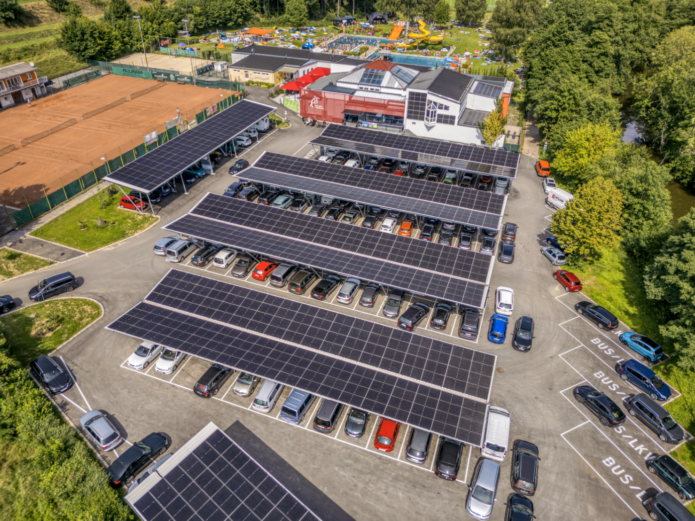 Luftaufnahme eines Parkplatzes mit Solarmodulen über den Autos. Das Gebiet ist von Bäumen und einem Tennisplatz umgeben.