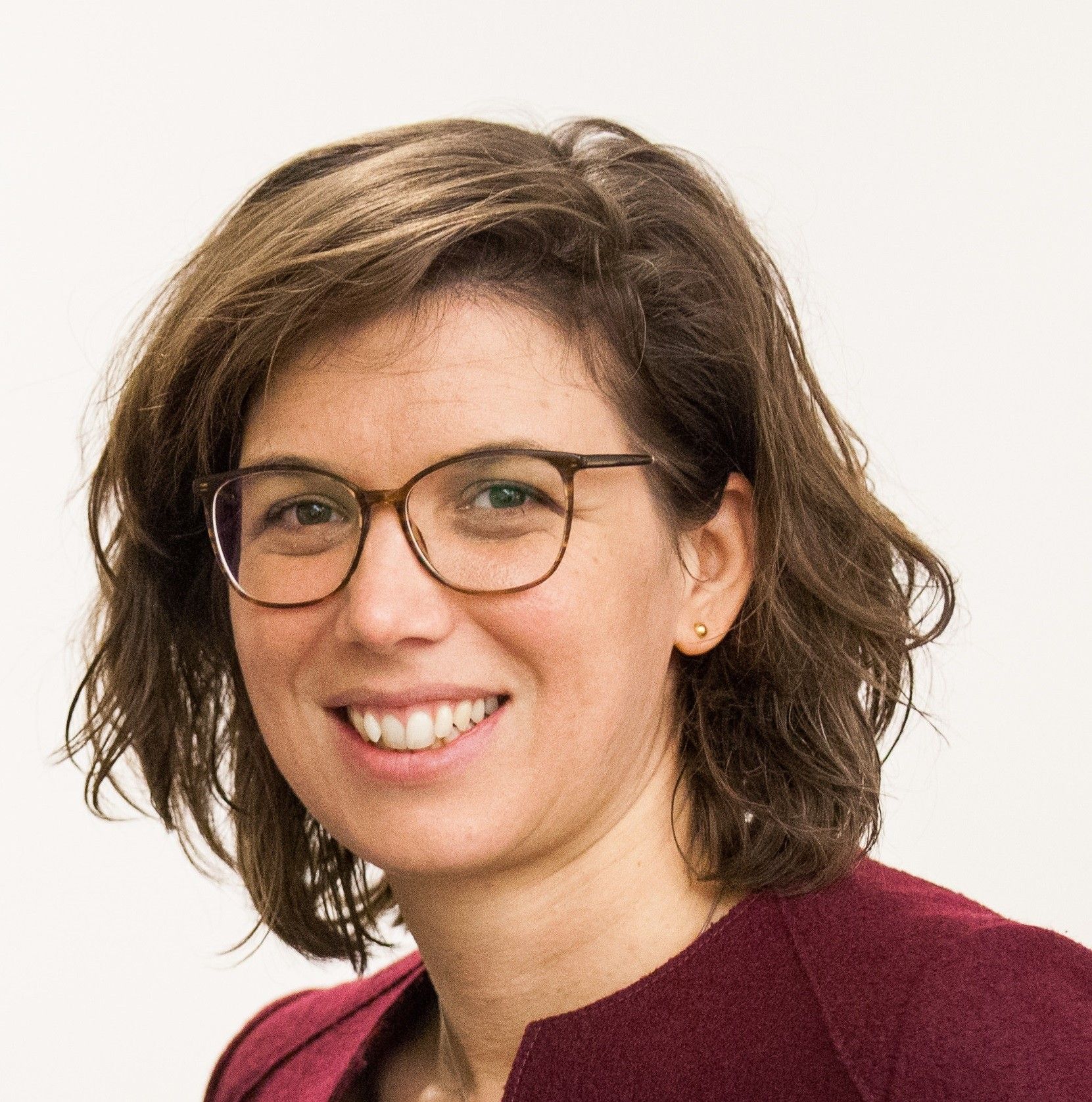 Eine Frau mit kurzen braunen Haaren und einer Brille lächelt für ein Porträt. Sie trägt ein weinrotes Hemd.